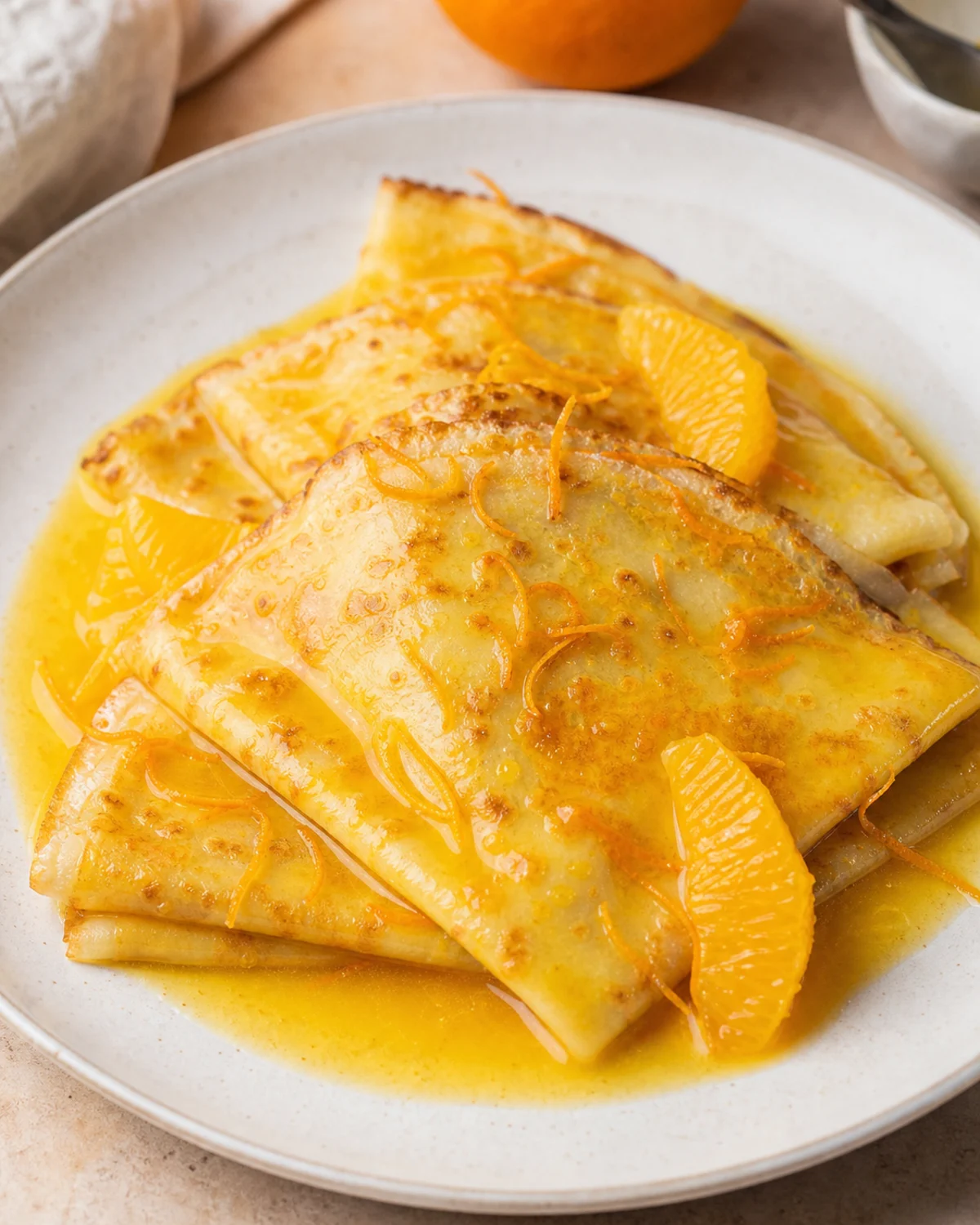 Crêpes Suzette pliées avec sauce orange brillante, zestes et segments d’orange