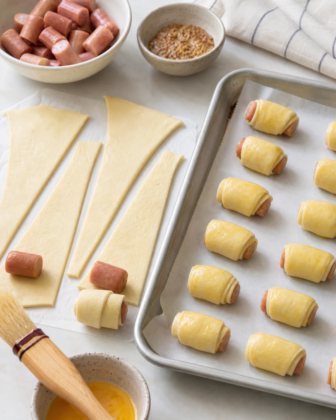 Préparation de feuilletés saucisse avec pâte découpée en triangles, petites saucisses coupées et bouchées dorées alignées sur une plaque avant cuisson