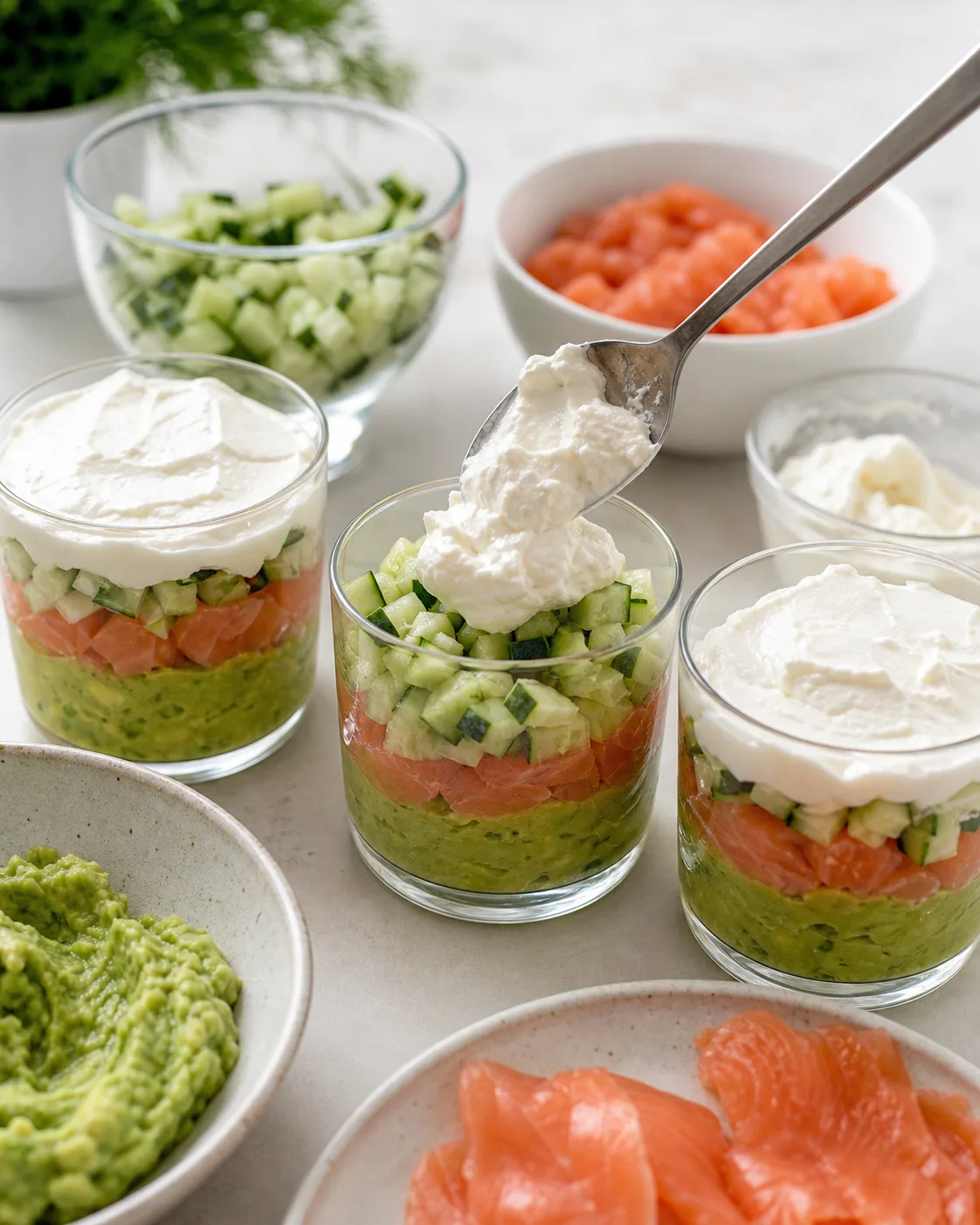 Montage de verrines saumon avocat avec base d’avocat, garniture saumon-concombre et crème au fromage frais dans petits verres transparents
